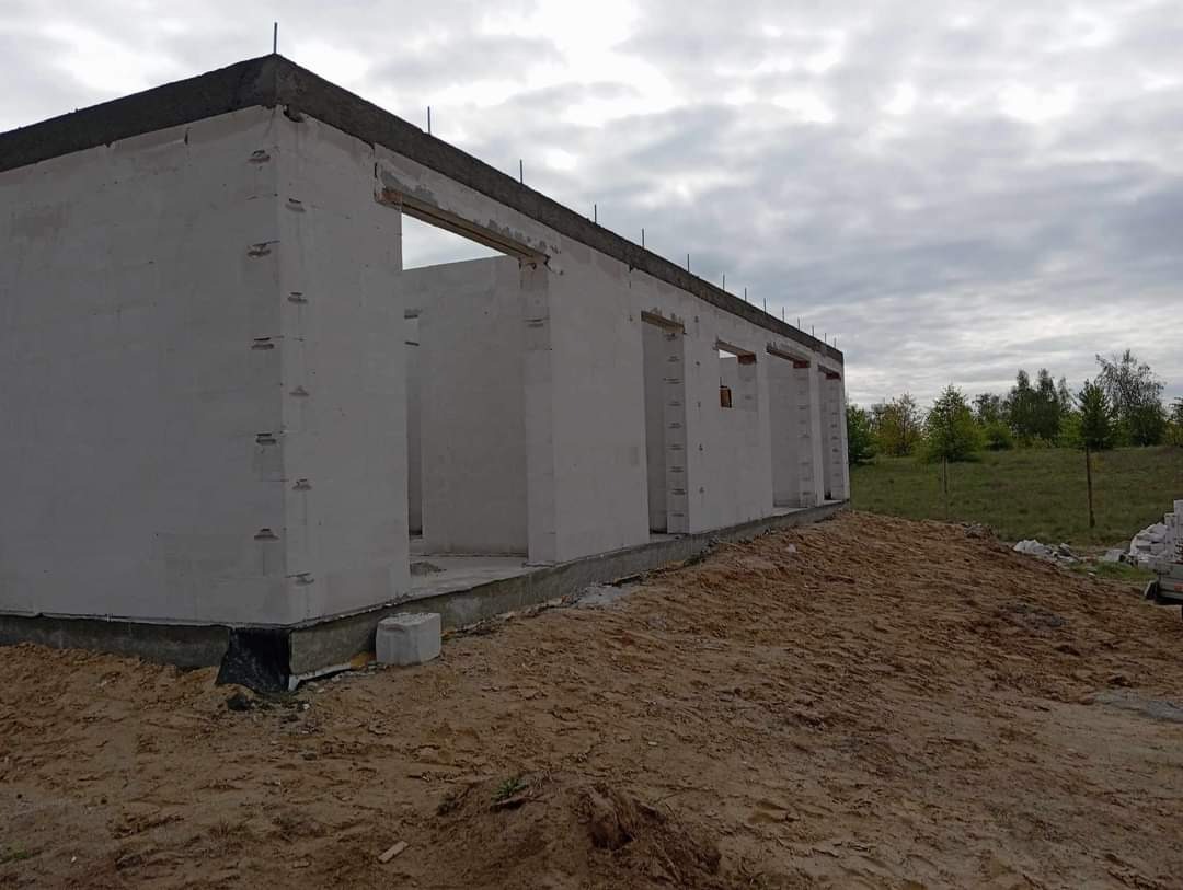 Surowy stan domu z betonu komórkowego z widocznymi otworami okiennymi i drzwiowymi, na etapie wznoszenia ścian, z ziemią i zielenią w tle pod pochmurnym niebem.