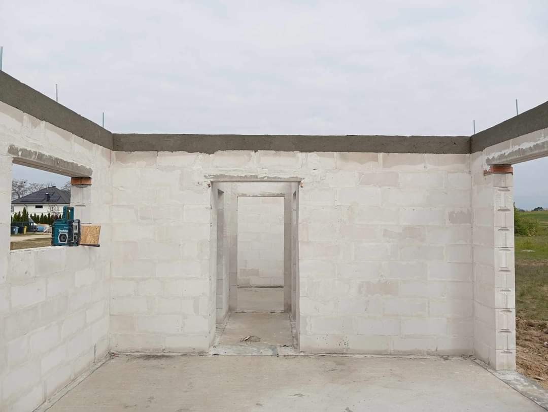 Wnętrze surowego, parterowego domu w trakcie budowy, ściany z białych bloczków, widoczne otwory okienne i drzwiowe, na parapecie poziomica laserowa.