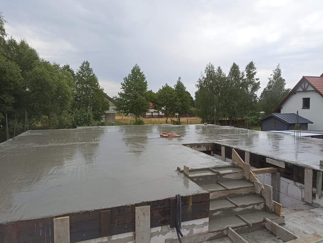 Świeżo wylany, mokry beton na fundamencie domu jednorodzinnego z widocznym szalunkiem schodów i wystającymi prętami zbrojeniowymi, tło stanowią drzewa i zabudowania.