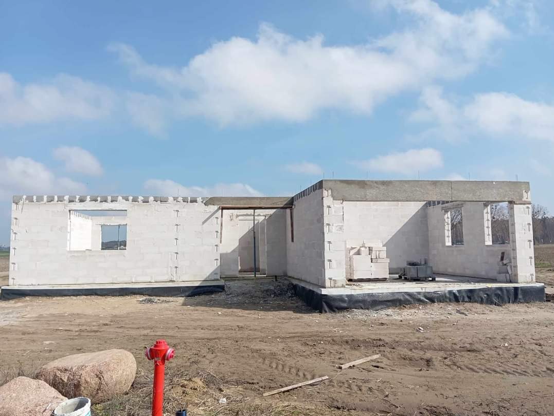 Niedokończony dom z białych bloczków, widoczne nadproża okienne, czerwony hydrant w pierwszym planie, błękitne niebo z chmurami w tle.