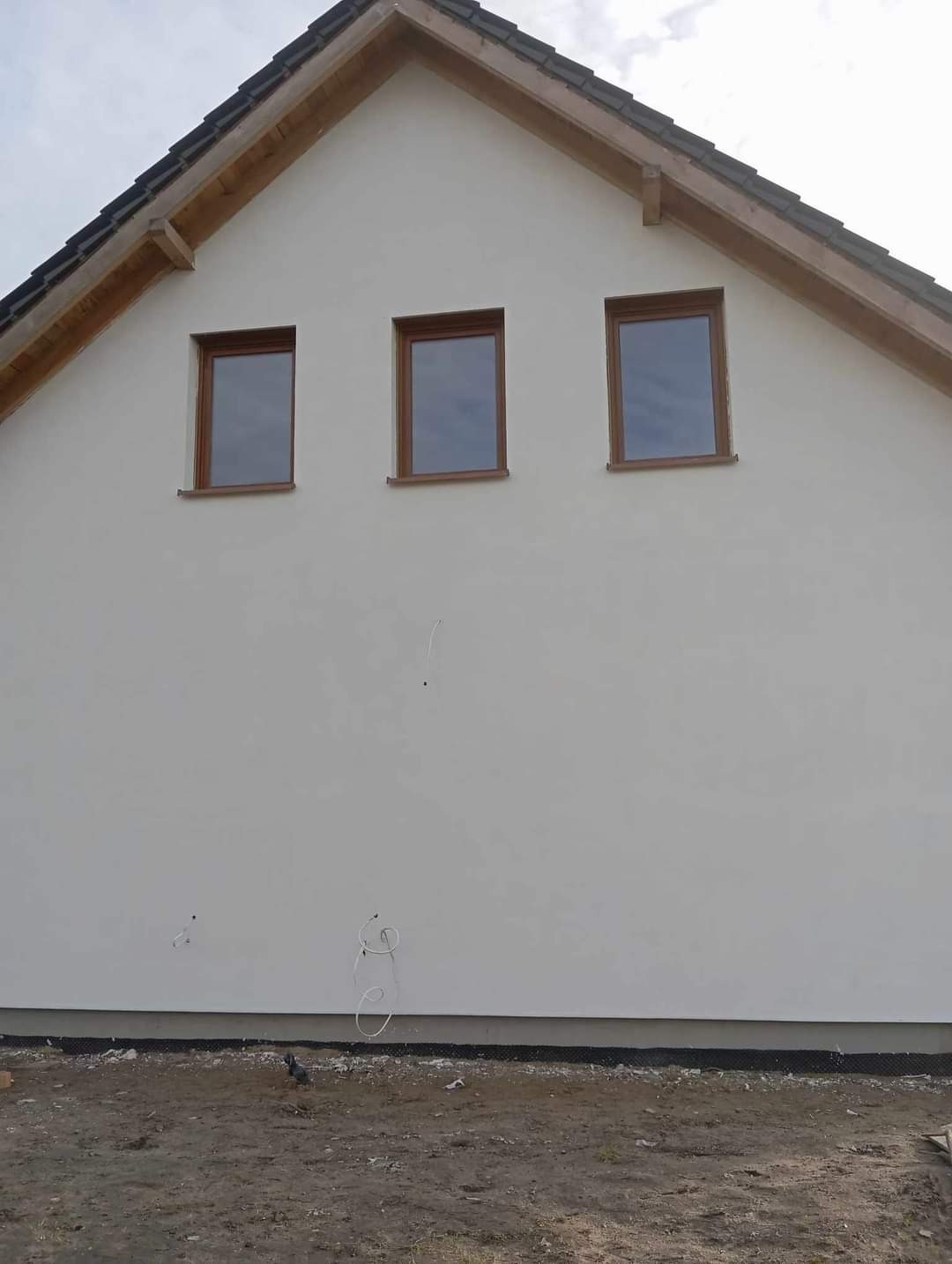 Boczny widok na nowo otynkowany dom z trzema oknami w drewnianych ramach, widoczne wystające kable i fragment gruntu.