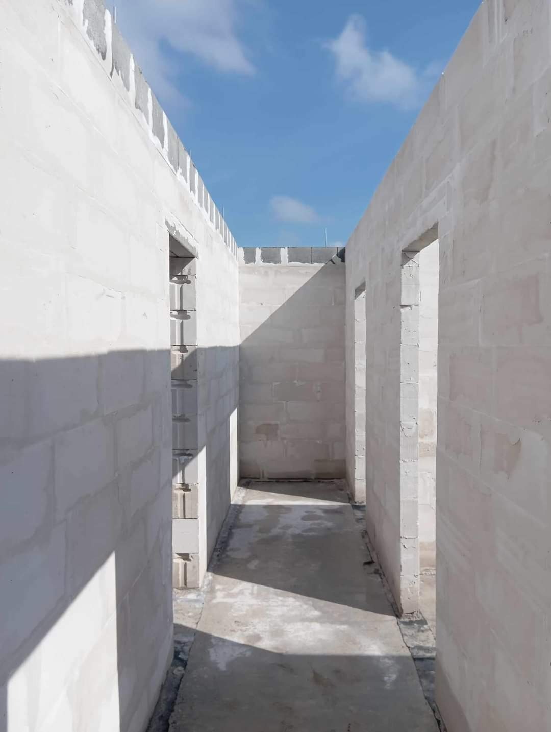 Wnętrze surowego, jasnego budynku w trakcie budowy z szarych bloczków, widoczne otwory drzwiowe i fragment nieba z chmurami.