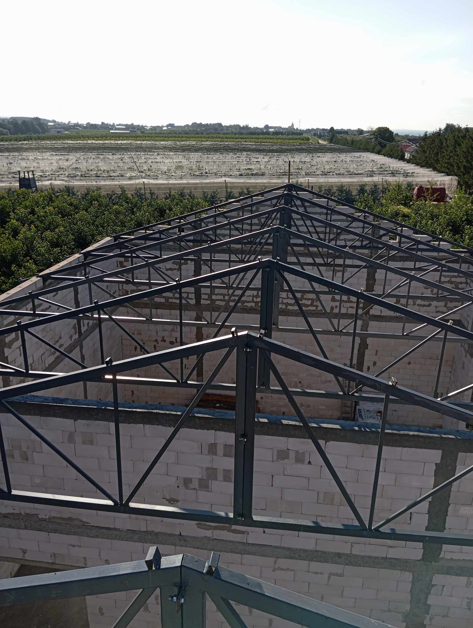 Wznoszona konstrukcja domu z białych bloczków, widok z góry na stalową konstrukcję dachu i odległe pola uprawne pod siatką.