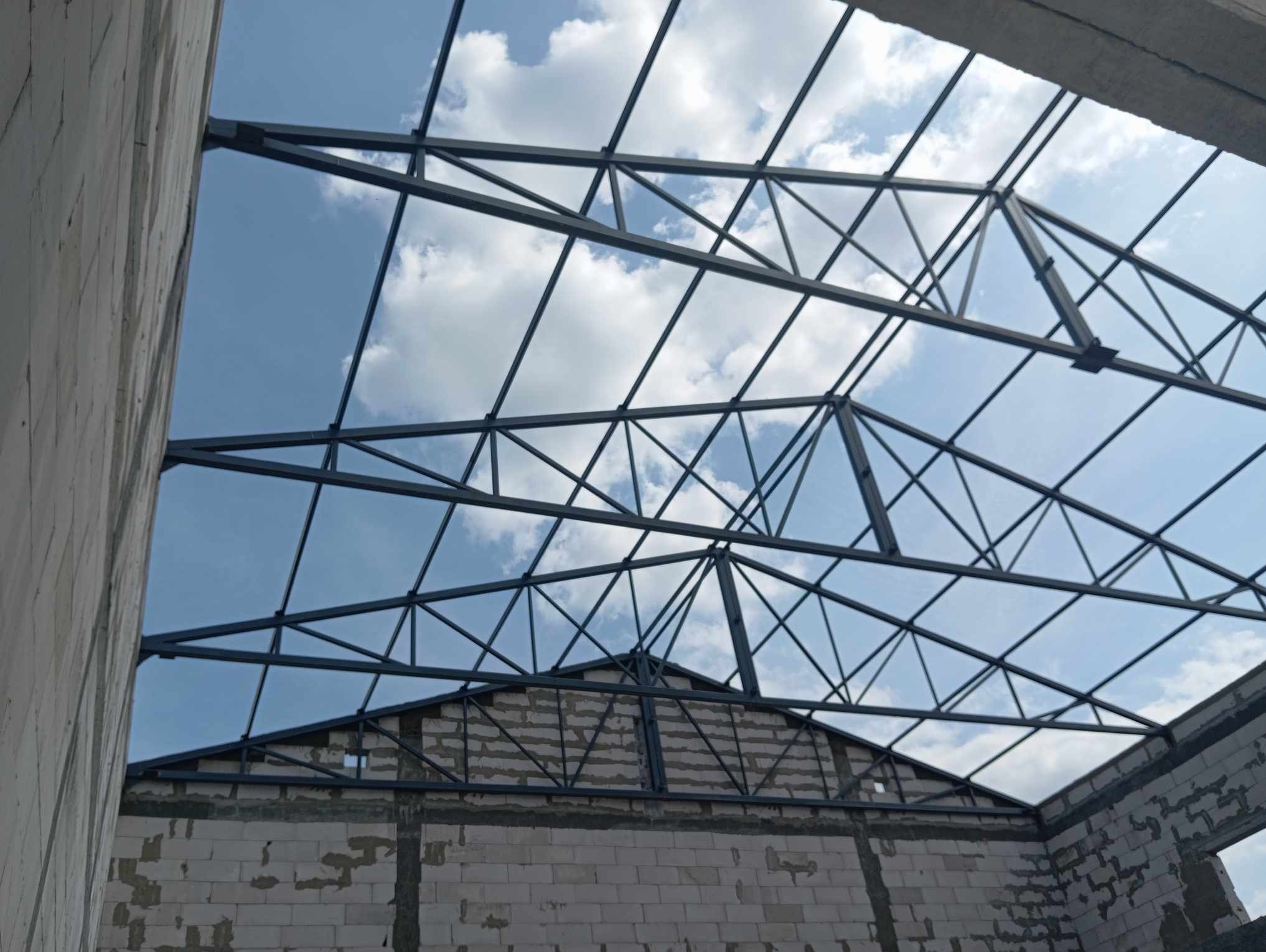 Konstrukcja stalowa dachu budynku w trakcie budowy, widok od wewnątrz na niebieskie niebo z chmurami, ściany z bloczków betonowych.