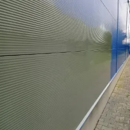 Szara, ryflowana elewacja budynku przemysłowego z niebieskimi akcentami, sfotografowana pod kątem, ukazująca fragment chodnika z kostki brukowej.