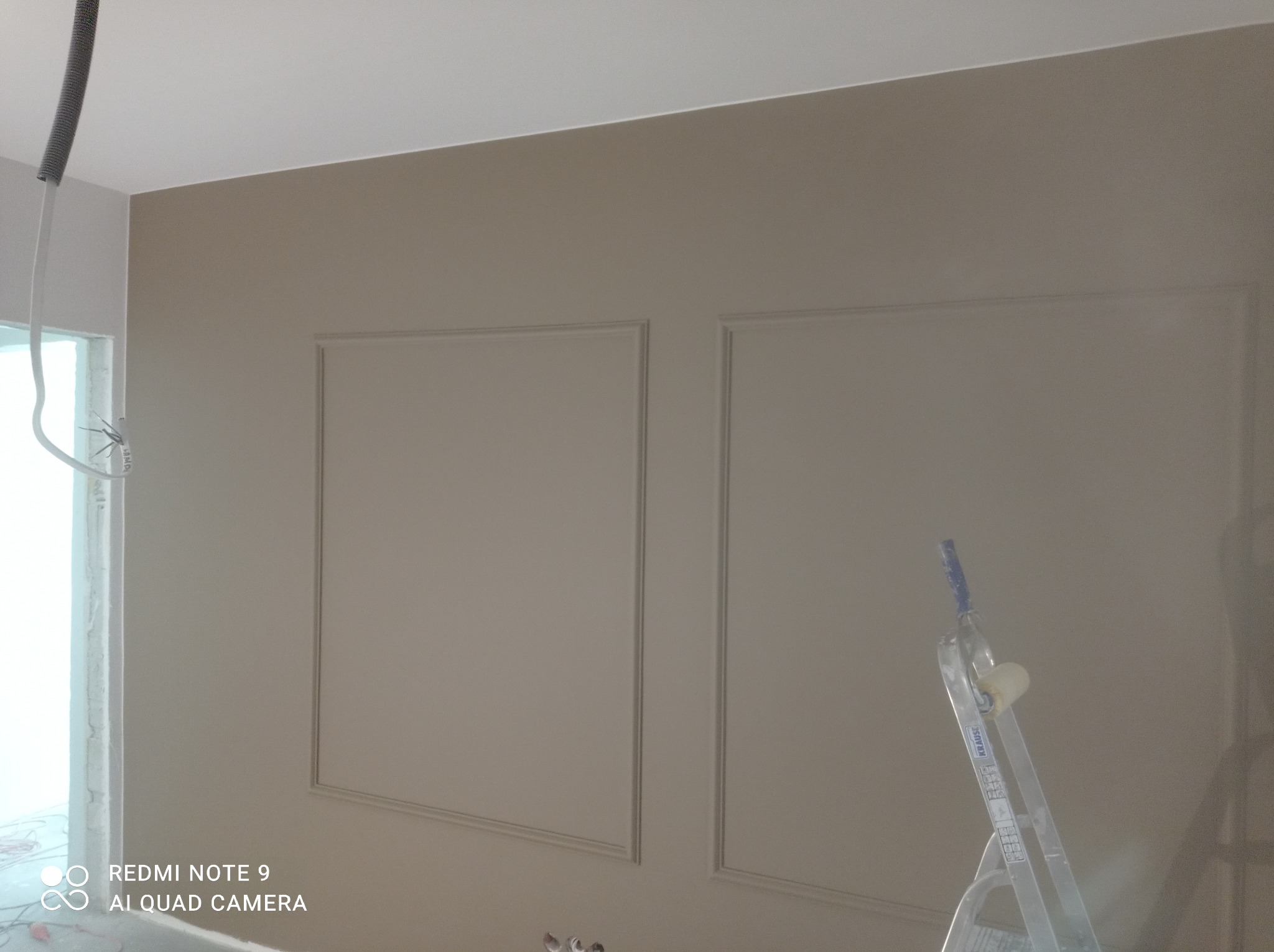 Ściana w odcieniach beżu z dekoracyjnymi ramkami, częściowo pomalowana, widoczna drabina malarska i przewody elektryczne wystające ze ściany.