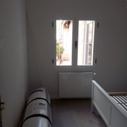 BART-BUD - Pokój w trakcie przygotowań do remontu: widoczne białe ściany, okno z widokiem na zewnątrz, grzejnik, częściowo złożone białe łóżko, zwinięty w folię materiał izolacyjny oraz białe tekstylia...