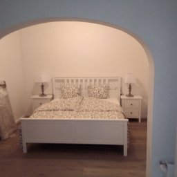 BART-BUD - Sypialnia z łóżkiem w stylu skandynawskim, widoczna przez łuk w ścianie, z podłogą w kolorze drewna i zapakowanymi elementami wystroju w rogu.