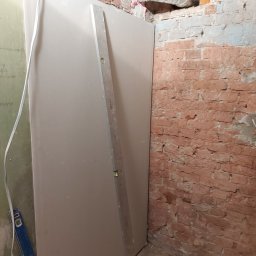 BART-BUD - Montaż płyty karton gips na ścianie z odsłoniętą cegłą, widoczny poziomica i fragment instalacji wentylacyjnej w otworze w ścianie.