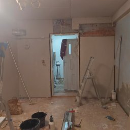 BART-BUD - Pokój w trakcie remontu: ściany wyłożone płytami kartonowo-gipsowymi, widoczne kable elektryczne, drabina, wiadra i narzędzia na podłodze, otwarte drzwi do kolejnego pomieszczenia.