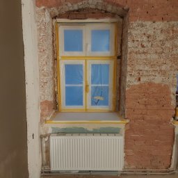 BART-BUD - Przygotowanie do prac wykończeniowych we Wrocławiu: okno zabezpieczone folią i taśmą malarską, fragment ściany z cegły z widocznymi śladami starego tynku, grzejnik. Widoczny etap prac przed...