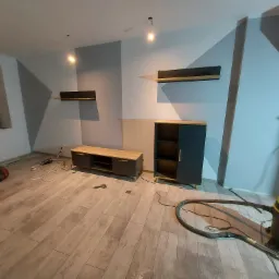 Wnętrze pokoju w trakcie wykończenia: ułożone panele podłogowe, częściowo pomalowane ściany w geometryczne wzory, zamontowane meble podwieszane i stojące, widoczny odkurzacz przemysłowy.