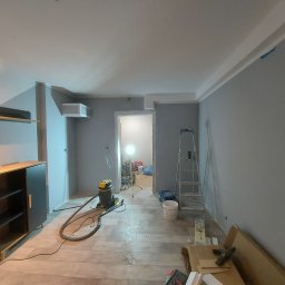 BART-BUD - Pokój w trakcie odświeżania: świeżo pomalowane szare ściany, drewnopodobne panele podłogowe, drabina oparta o ścianę, odkurzacz przemysłowy i resztki materiałów budowlanych na podłodze.