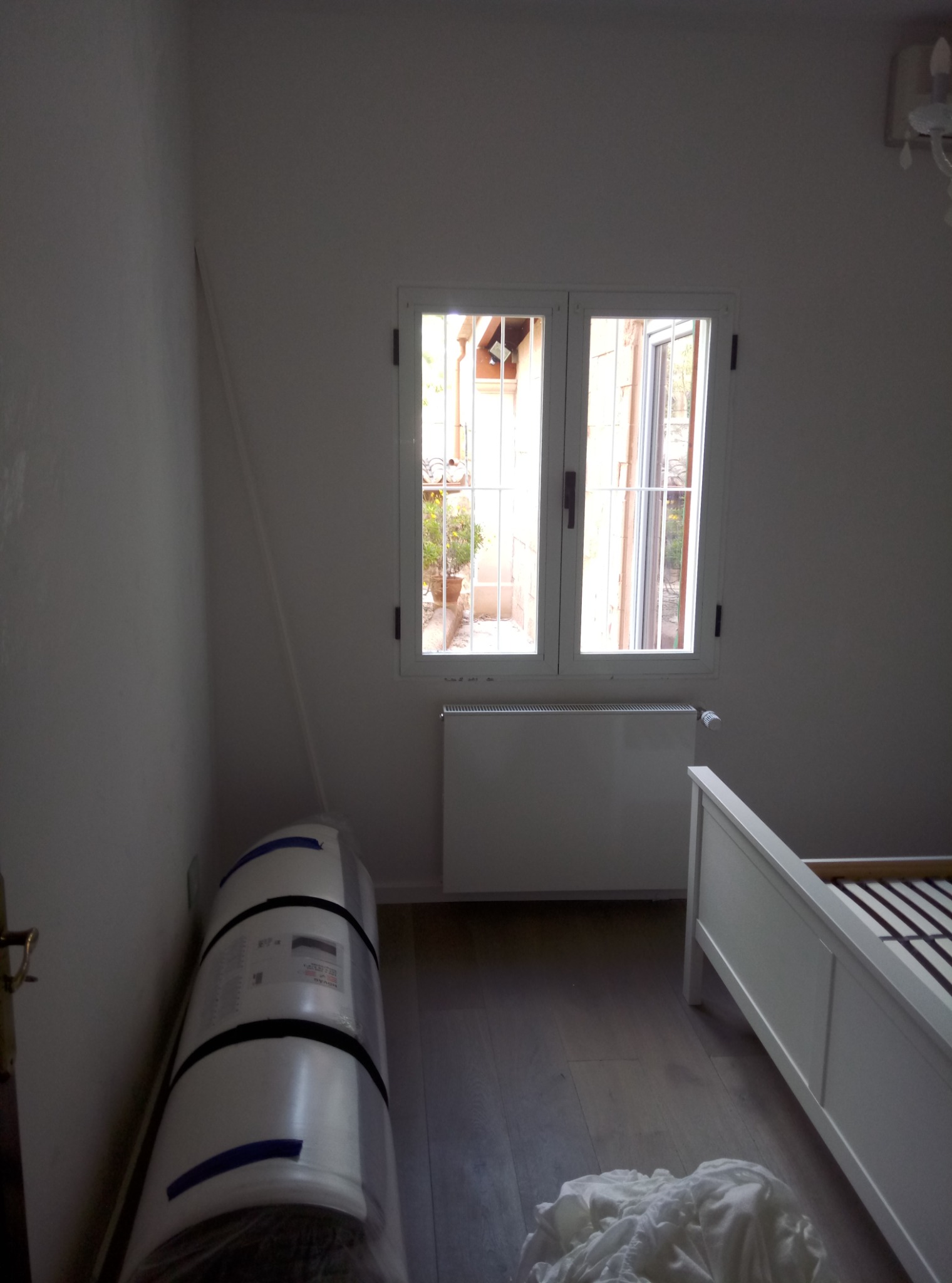 Pokój w trakcie przygotowań do remontu: widoczne białe ściany, okno z widokiem na zewnątrz, grzejnik, częściowo złożone białe łóżko, zwinięty w folię materiał izolacyjny oraz białe tekstylia...