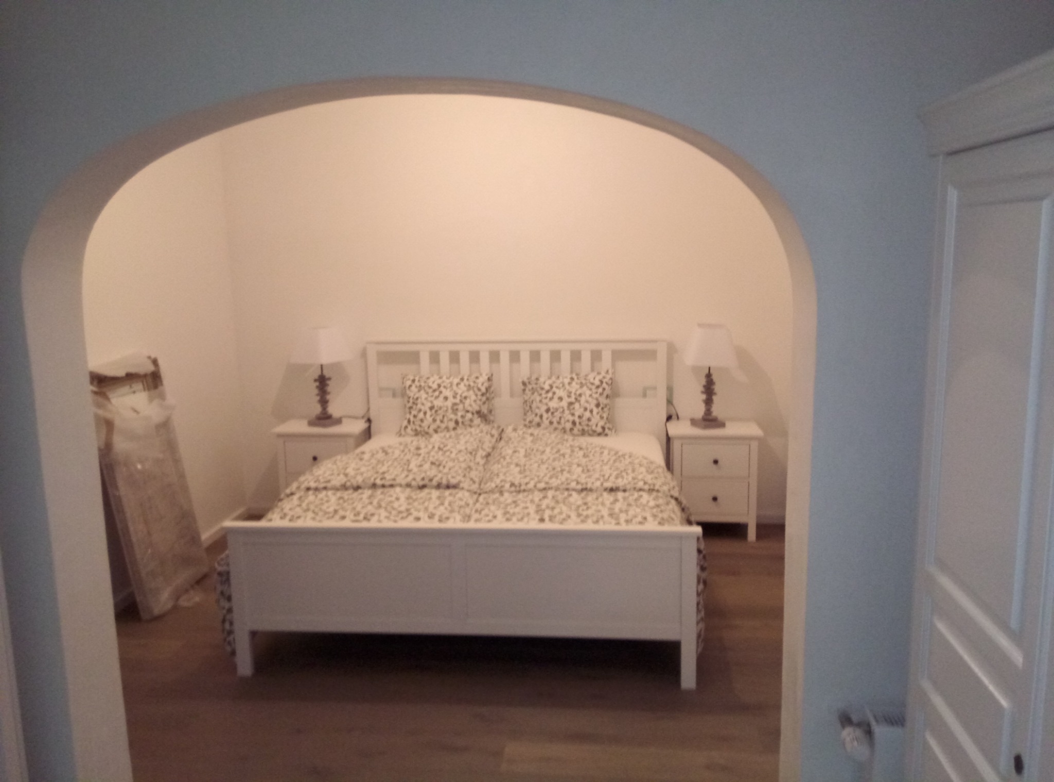 Sypialnia z łóżkiem w stylu skandynawskim, widoczna przez łuk w ścianie, z podłogą w kolorze drewna i zapakowanymi elementami wystroju w rogu.