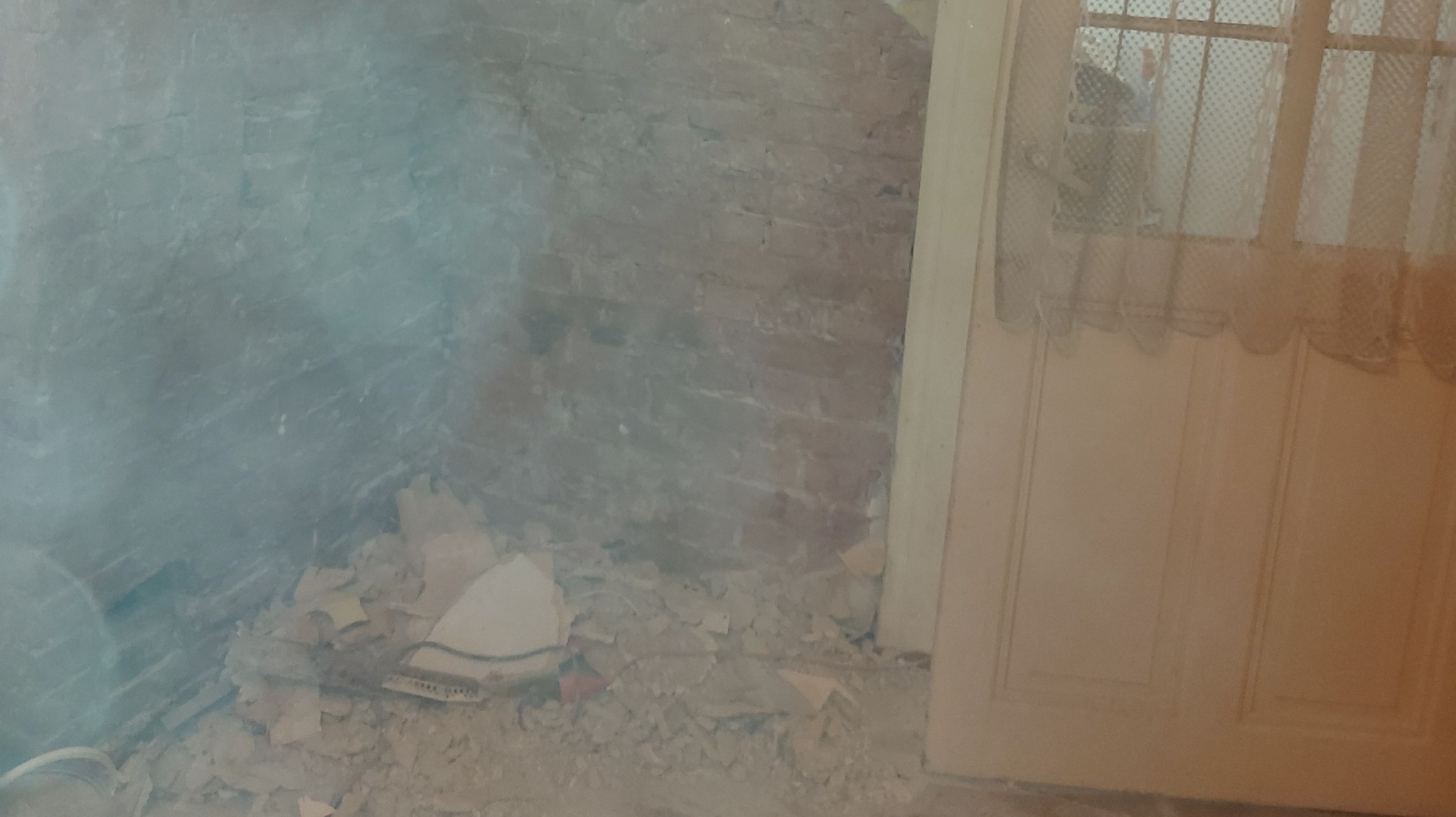 Fragment ściany z odsłoniętą cegłą i resztkami gruzu, widoczny fragment drzwi z firanką w tle.