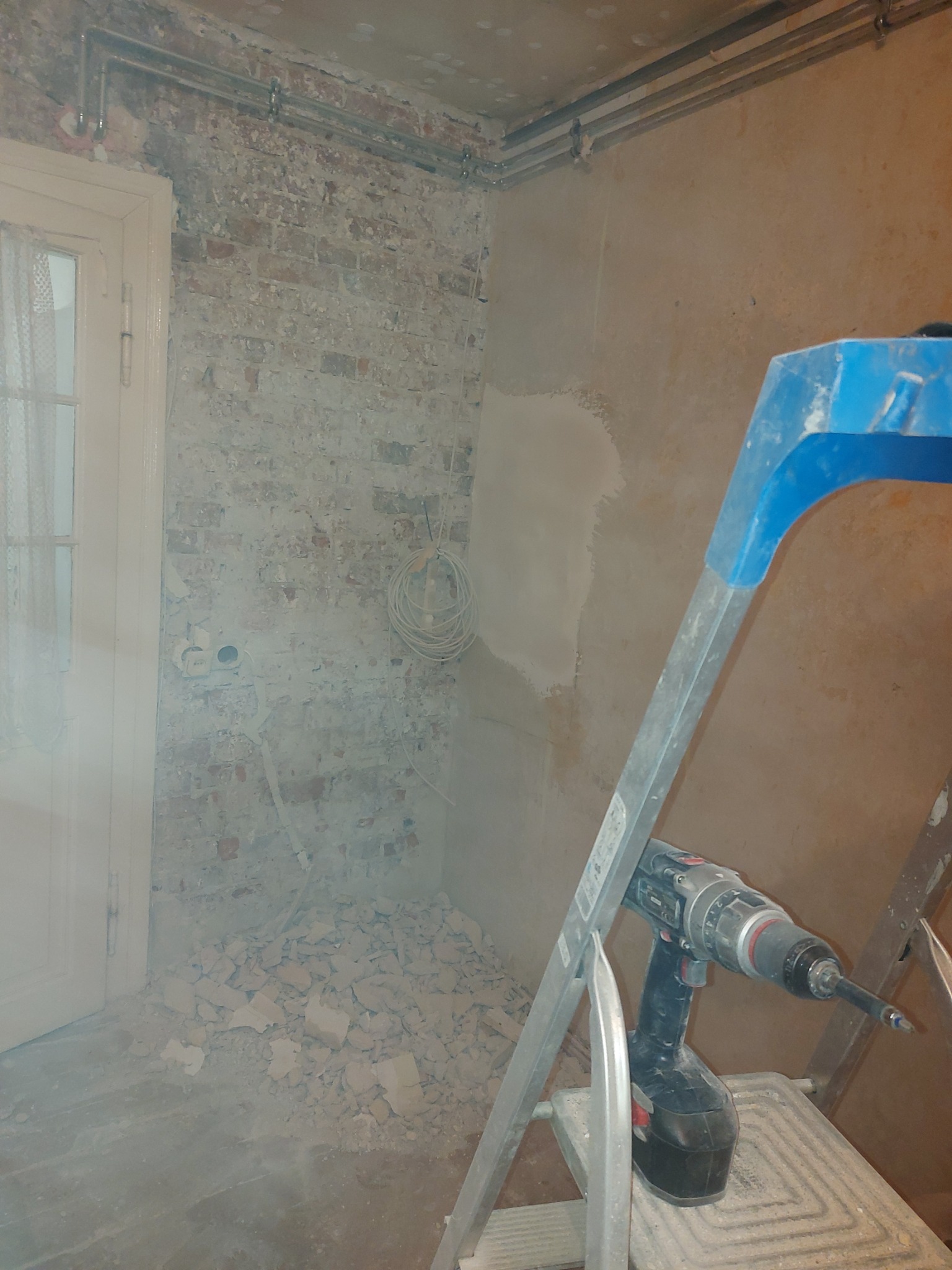 Wnętrze w trakcie remontu: fragment ściany z odsłoniętą cegłą, fragment otynkowanej ściany, przewody elektryczne, gruz na podłodze i drabina z wiertarką.