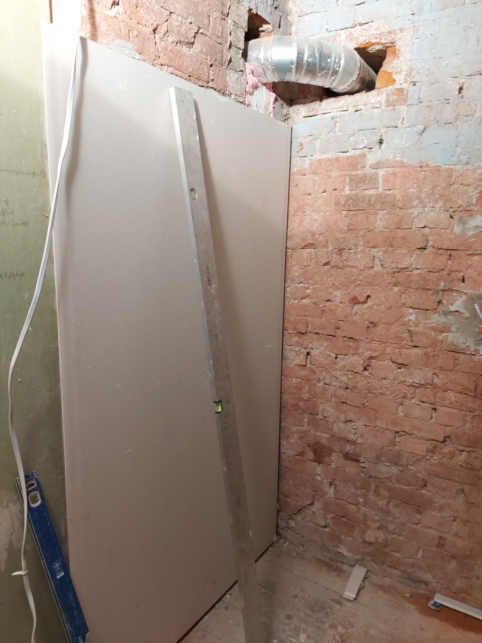 Montaż płyty karton gips na ścianie z odsłoniętą cegłą, widoczny poziomica i fragment instalacji wentylacyjnej w otworze w ścianie.