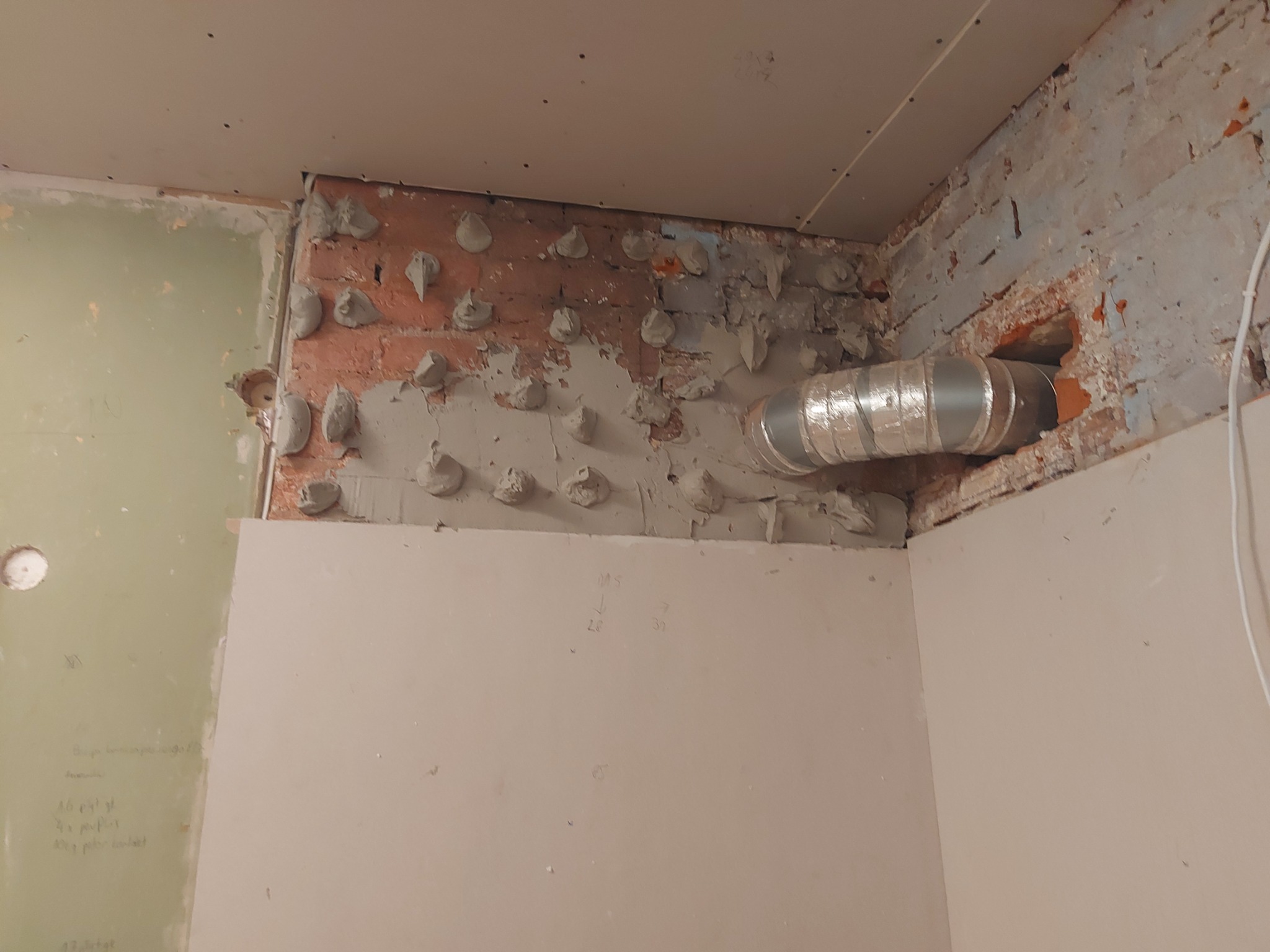 Fragment ściany z cegły z widocznymi punktami mocowania, częściowo zakrytej płytą kartonowo-gipsową oraz rurą wentylacyjną, ukazujący etap prac wykończeniowych.