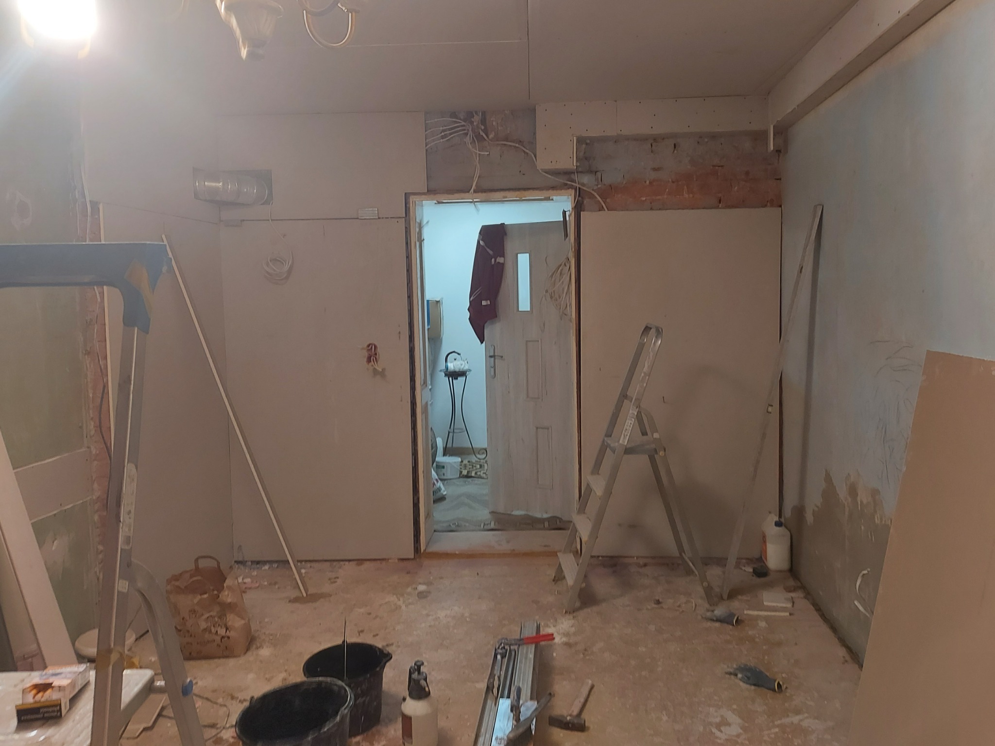 Pokój w trakcie remontu: ściany wyłożone płytami kartonowo-gipsowymi, widoczne kable elektryczne, drabina, wiadra i narzędzia na podłodze, otwarte drzwi do kolejnego pomieszczenia.