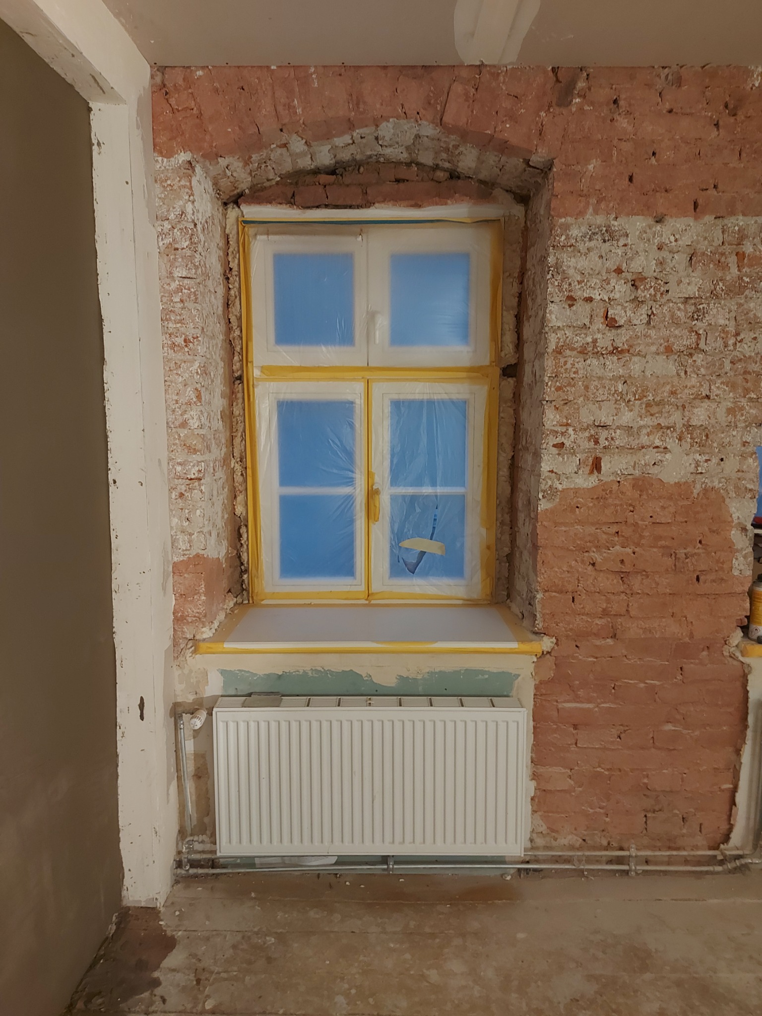 Przygotowanie do prac wykończeniowych we Wrocławiu: okno zabezpieczone folią i taśmą malarską, fragment ściany z cegły z widocznymi śladami starego tynku, grzejnik. Widoczny etap prac przed...