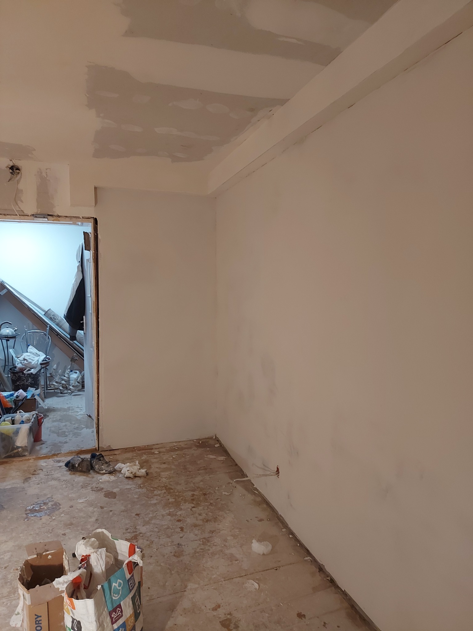 Pokój w trakcie prac wykończeniowych: świeżo pomalowana ściana w odcieniach bieli, widoczne poprawki na suficie, podłoga częściowo zakryta folią i resztkami materiałów budowlanych, torby z zaprawą...