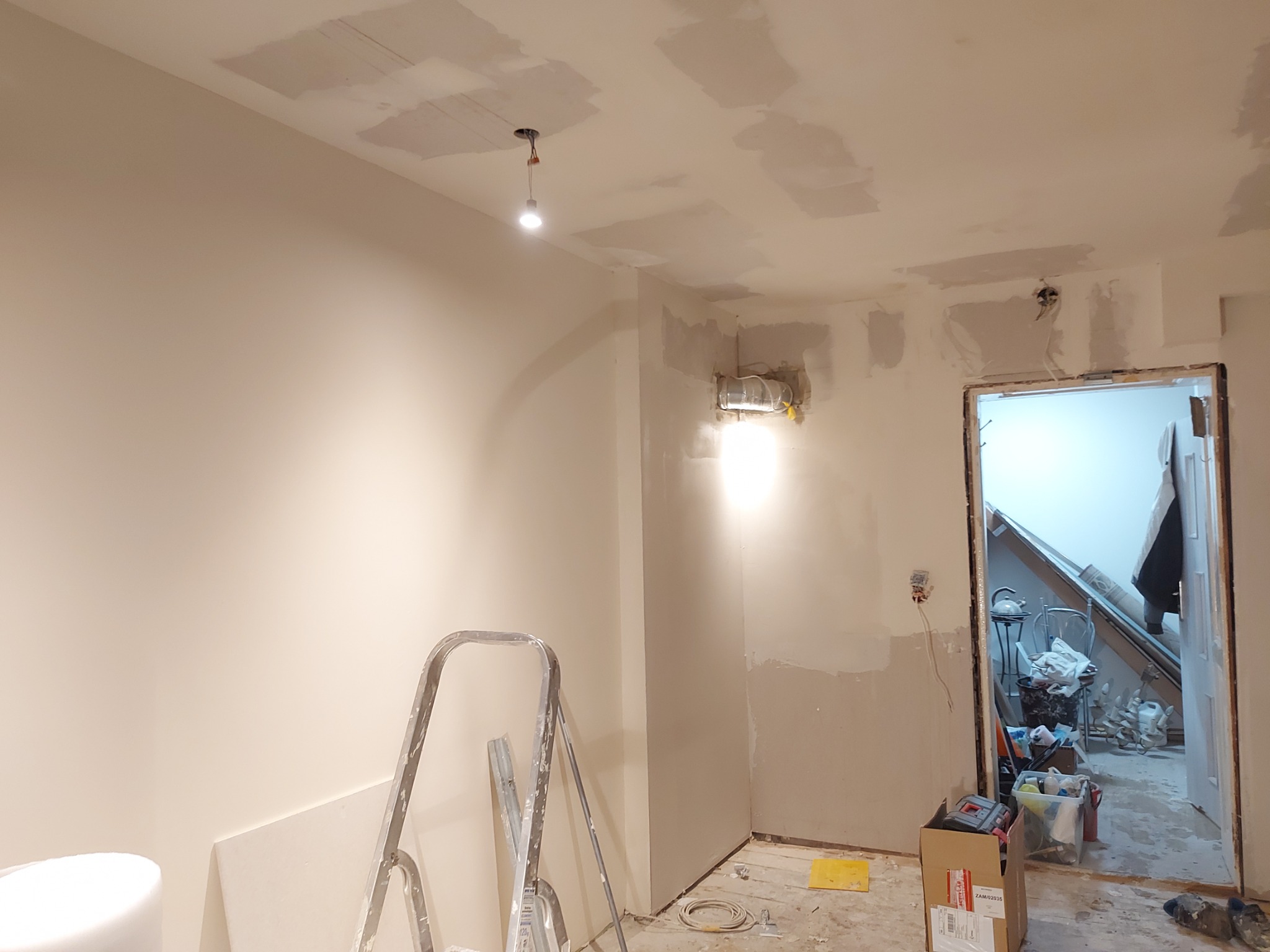 Wnętrze w trakcie remontu: ściany przygotowane do malowania, widoczne ślady po szpachlowaniu na suficie i ścianach, drabina malarska, niezabezpieczona instalacja elektryczna, bałagan w tle.