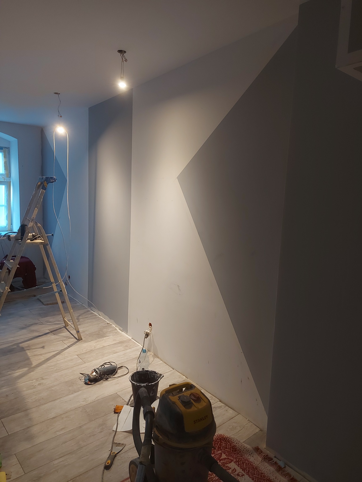 Pokój w trakcie remontu: świeżo pomalowane szare ściany z geometrycznym wzorem, drewniana drabina, szlifierka kątowa, wiadro z farbą i odkurzacz przemysłowy na panelach podłogowych.