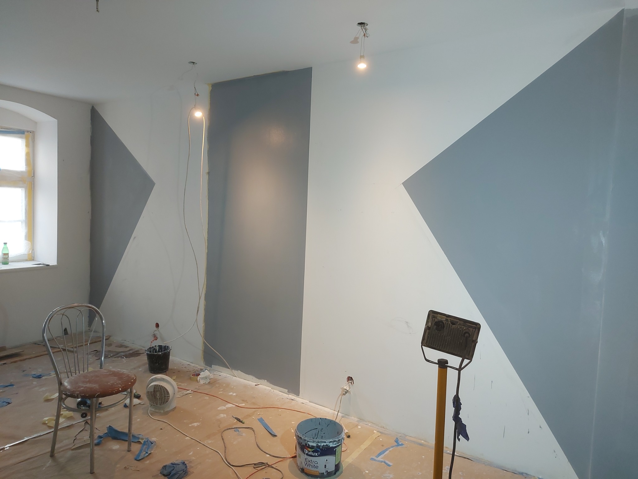 Wnętrze pokoju w trakcie malowania z geometrycznym wzorem w odcieniach szarości i bieli, widoczne wiadro z farbą Dulux Extra White, metalowe krzesło i halogen na statywie.