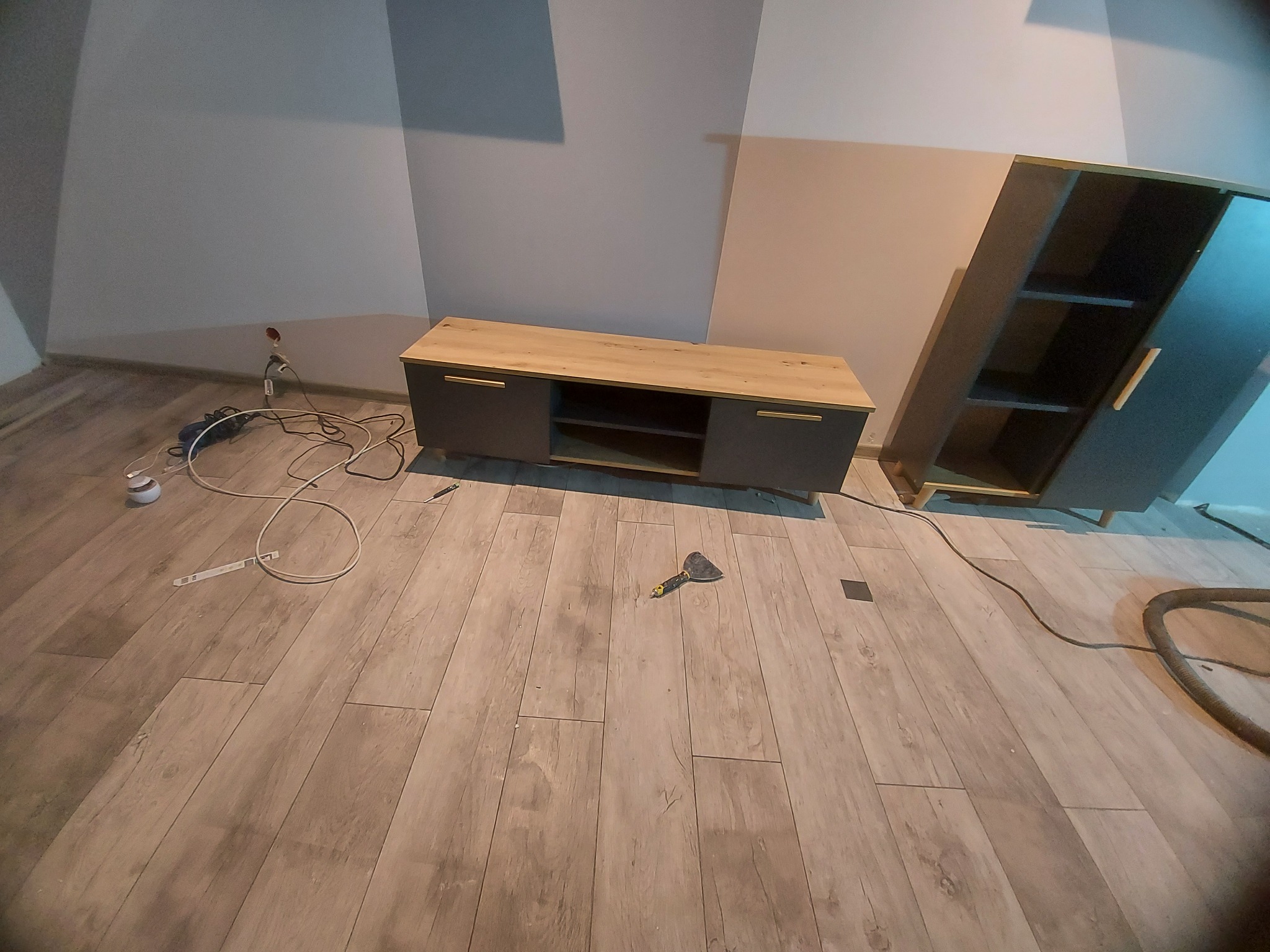 Pokój po remoncie z nową podłogą z paneli, widoczne listwy przypodłogowe, szafka RTV i szafa oraz narzędzie do układania paneli na podłodze.