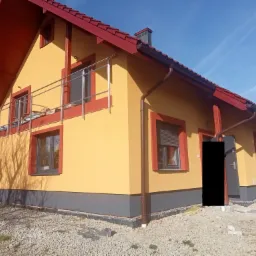 Żółty dom jednorodzinny z czerwonym dachem i szarą podmurówką, widoczny z perspektywy ogrodu przed domem.