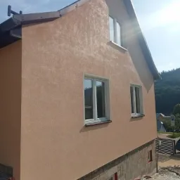 Świeżo otynkowana elewacja domu jednorodzinnego w odcieniach beżu, z widocznymi oknami z białymi ramami i fragmentem kamiennej podmurówki, na tle zieleni i błękitnego nieba.