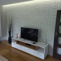 Ściana z białej cegły dekoracyjnej z podświetleniem LED, biała szafka RTV z drewnianymi akcentami, drewniana podłoga, czarny wazon z suchymi gałązkami i telewizor.