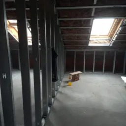 Poddasze w trakcie adaptacji z widoczną konstrukcją z profili stalowych, ociepleniem wełną mineralną i oknem dachowym; na podłodze prowizoryczny stół z cegieł i butelka z żółtym płynem.