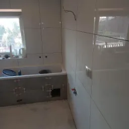 Białe, błyszczące płytki ścienne w trakcie montażu w łazience, widoczne krzyżyki dystansowe, przygotowana obudowa wanny z szarych płytek, wystające przewody elektryczne i rury wodne.