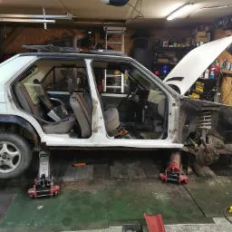 Biały samochód osobowy, podniesiony na lewarkach w warsztacie, z otwartą maską i usuniętymi elementami karoserii, ukazujący postęp prac renowacyjnych.