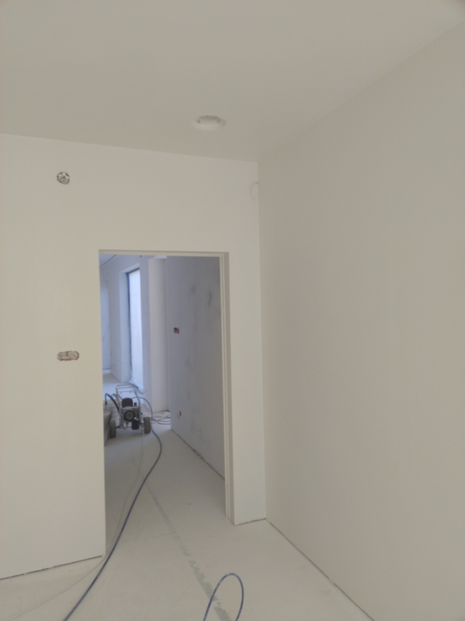 Wnętrze w trakcie prac wykończeniowych, widoczna ściana po nałożeniu gładzi, w tle agregat malarski i przygotowane podłoże.