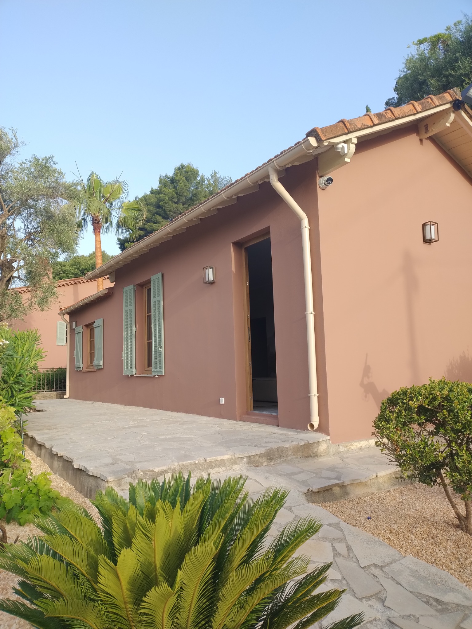 Odnowiony domek jednorodzinny z elewacją w kolorze terakoty, jasnymi okiennicami i systemem rynnowym, widoczne rośliny tropikalne w ogrodzie.