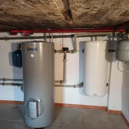 Dwa zbiorniki ciepłej wody użytkowej, Ferroli i Galmet, z widocznymi przyłączami hydraulicznymi i zaworami w piwnicy z surowym betonowym sufitem.