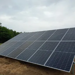 Rząd paneli słonecznych na gruncie, widoczne mocowania, pochmurne niebo w tle.