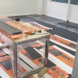 Przygotowania do montażu paneli: rozłożone opakowania paneli marki b.design na podłodze z zaznaczonymi liniami montażowymi, stół owinięty folią z narzędziami pomiarowymi na wierzchu, widok na okno...