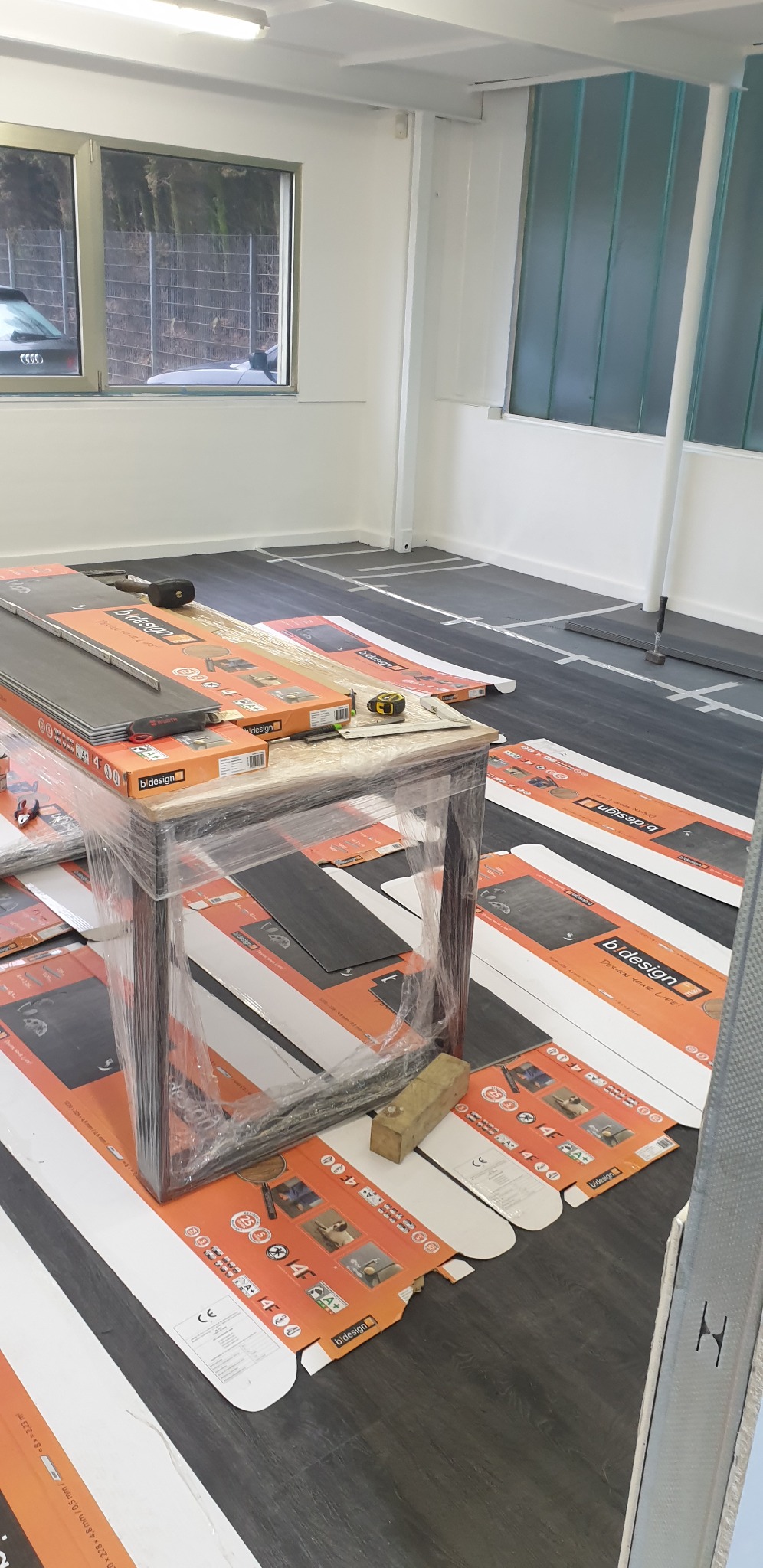 Przygotowania do montażu paneli: rozłożone opakowania paneli marki b.design na podłodze z zaznaczonymi liniami montażowymi, stół owinięty folią z narzędziami pomiarowymi na wierzchu, widok na okno...