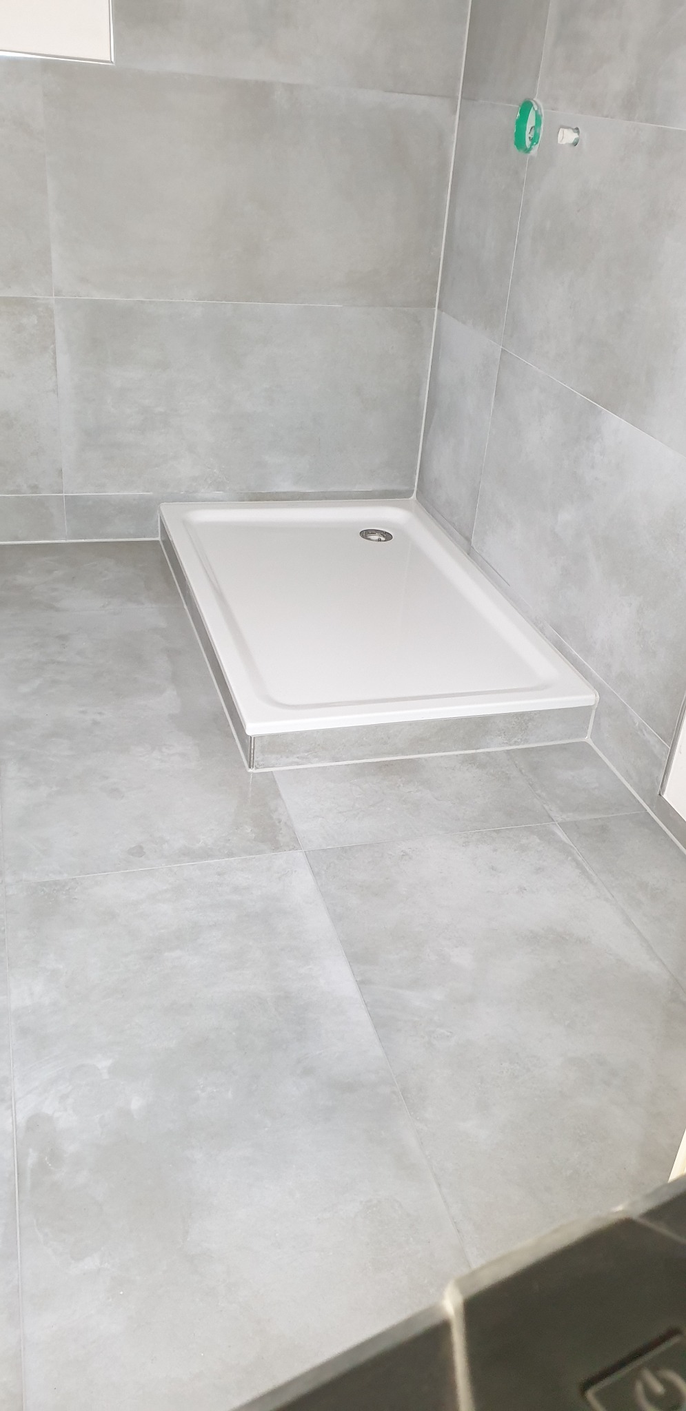 Nowoczesny, biały brodzik z odpływem wkomponowany w szare, matowe płytki podłogowe i ścienne, widoczny fragment instalacji wodnej.