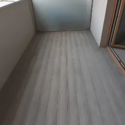 Wykończony balkon z szarymi panelami podłogowymi ułożonymi wzdłuż, częściowo przeszkloną balustradą i widokiem na budynek mieszkalny w tle.