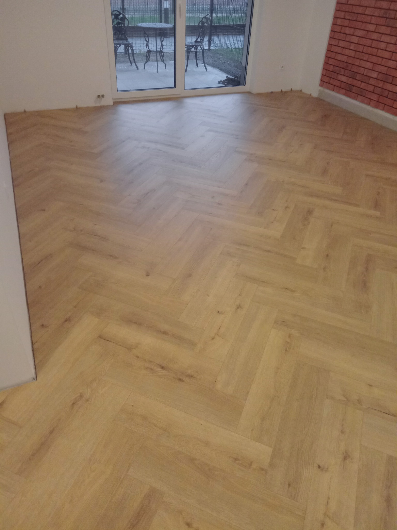 Jasna podłoga ułożona w jodełkę francuską, widoczne listwy przypodłogowe i okno balkonowe z widokiem na stolik i krzesła ogrodowe.