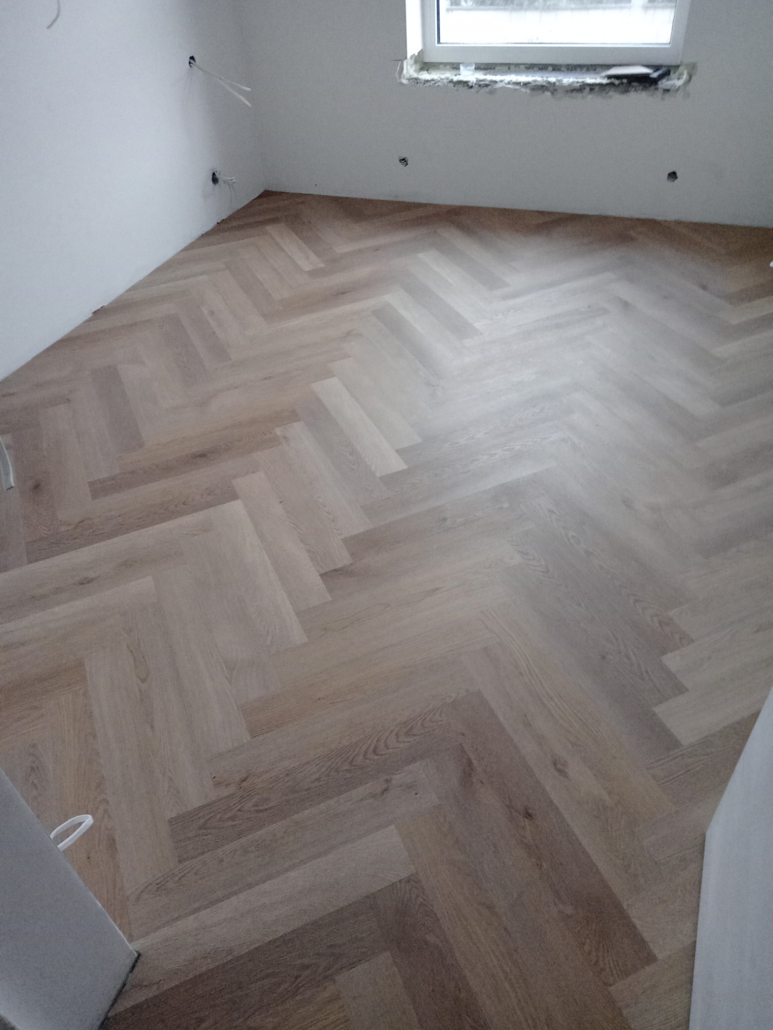 Jasny, drewnopodobny wzór ułożony w jodełkę na podłodze w surowym, białym pomieszczeniu z widocznymi przewodami elektrycznymi i niedokończonym oknem.