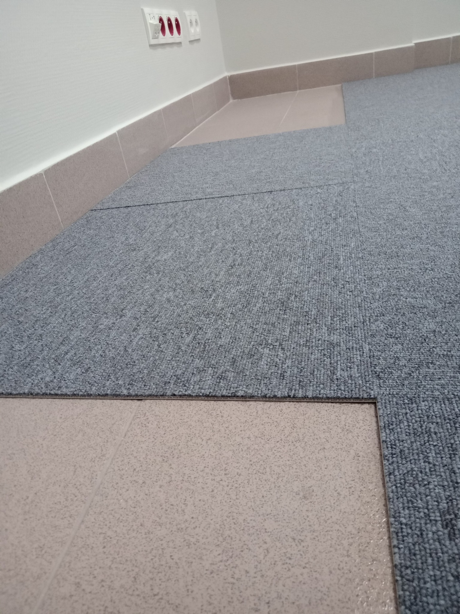 Szare kwadratowe płytki wykładzinowe układane na podłodze z jasnych płytek ceramicznych, widoczne gniazdka elektryczne na ścianie.