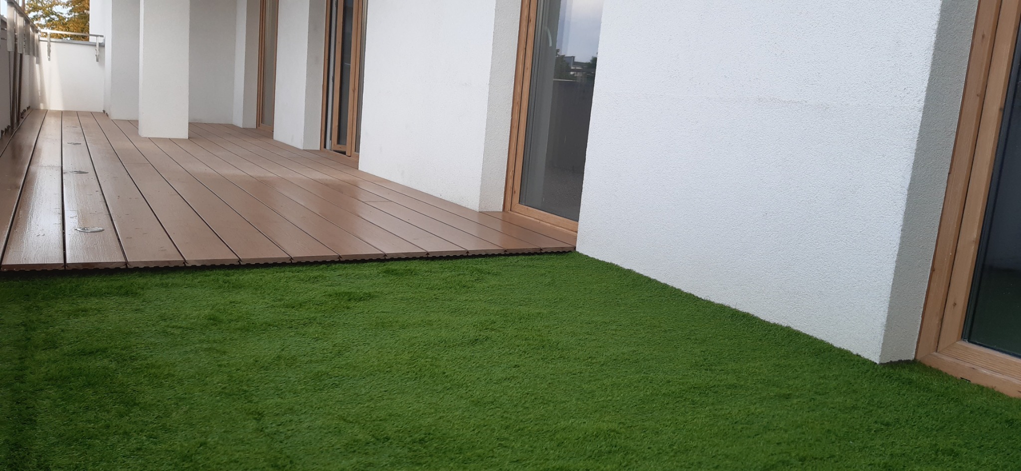 Kąt ujęcia balkonu z brązową podłogą z desek kompozytowych i sztuczną trawą przylegającą do białej ściany budynku z drewnianą ramą okienną.