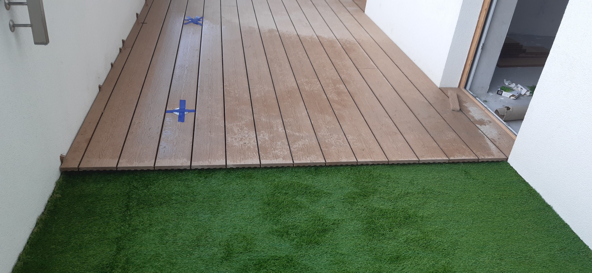 Balkon z brązowych paneli kompozytowych i sztucznej trawy, zabezpieczony niebieską taśmą malarską w trakcie montażu.