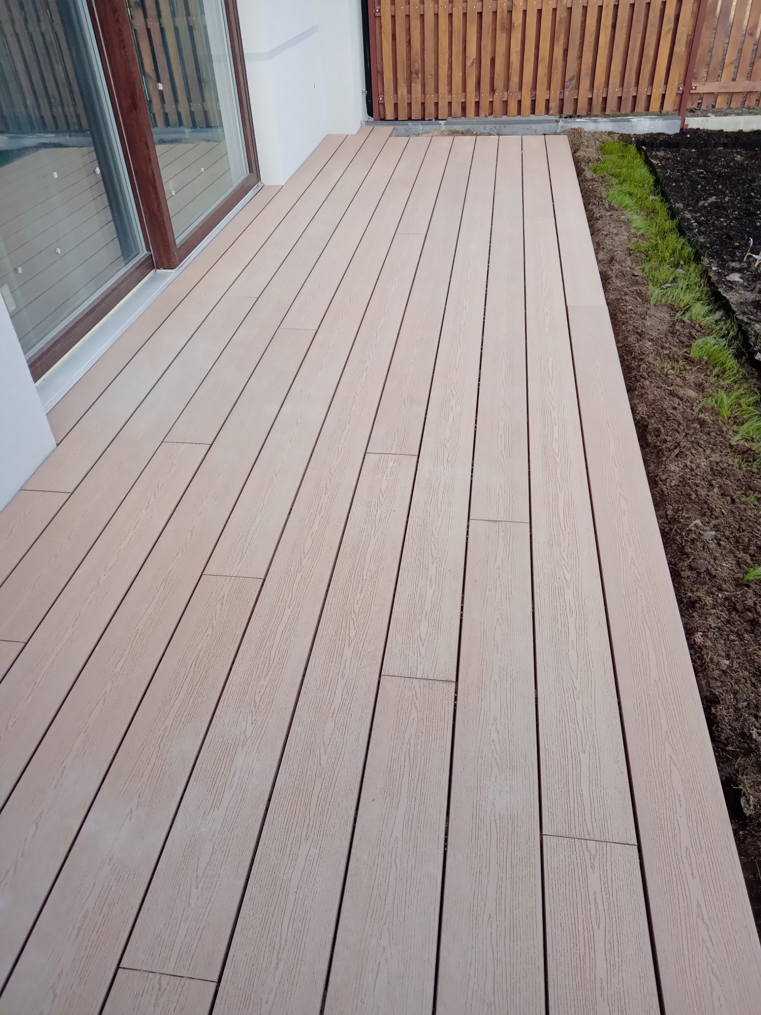 Beżowy taras z desek kompozytowych przylegający do budynku z dużym oknem, widoczna ziemia i drewniane ogrodzenie w tle.